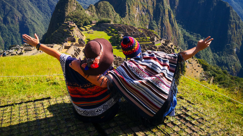 Two people dressed in traditional ponchos spread their arms toward Machu Picchu, Peru