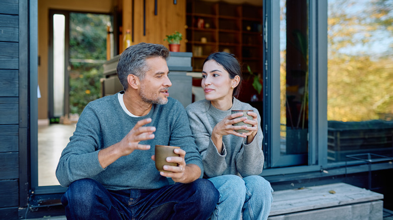 A man and woman sit on a house's porch with mugs of coffee