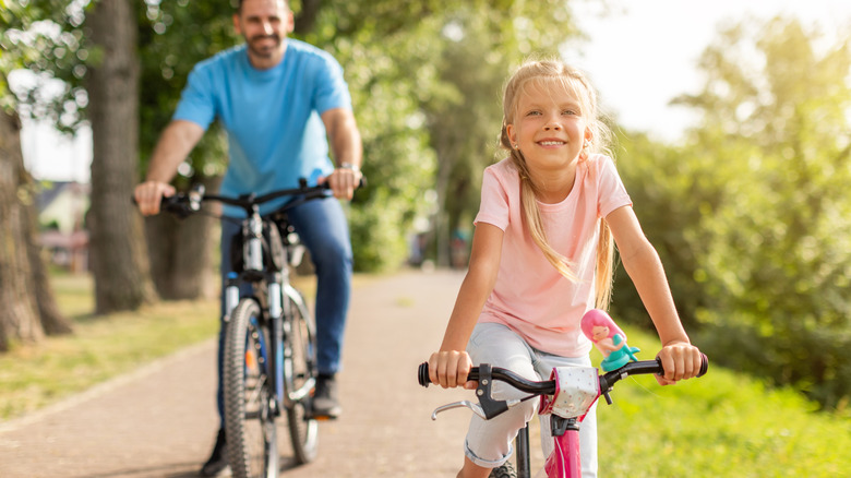 A father and daughter ride bicycles on a paved path