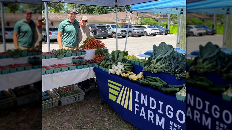 Two men pose and smile next to Indigo Farm tent where fruit and vegetables are on display