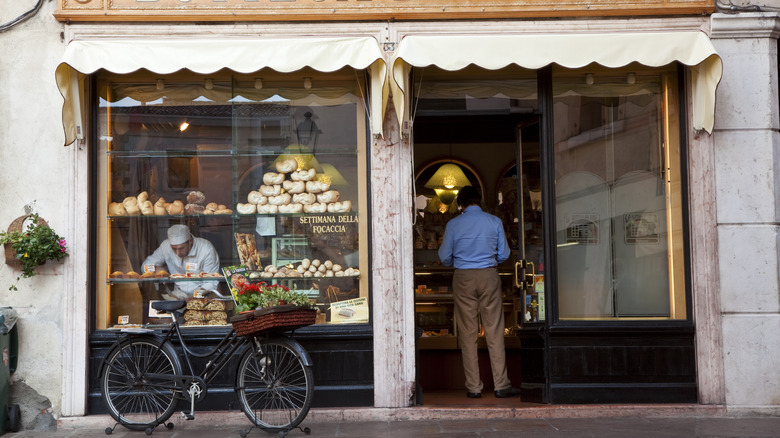 A bike with flowers in a basket is parked in front of an Italian bakery, where a chef arranges pastries in the window cast