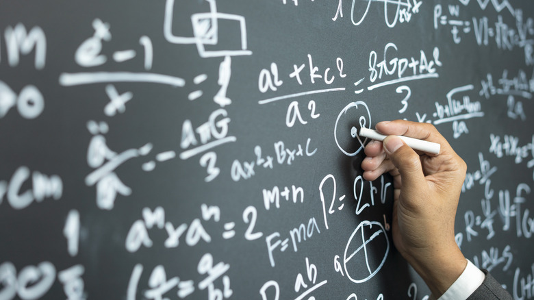 A hand doing equations with white chalk on a black chalkboard
