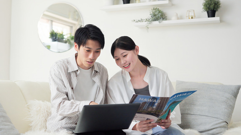 Happy couple plans vacation on couch using brochure and laptop.
