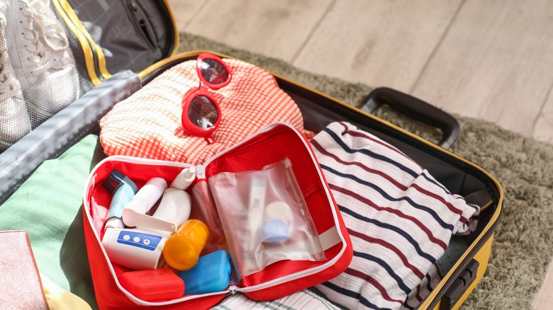 Suitcase with open first aid kit, stylish clothes on floor in room