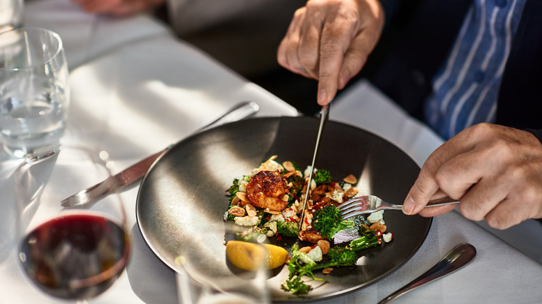 Hands holding dining utensils over a plate of food on an elegant table