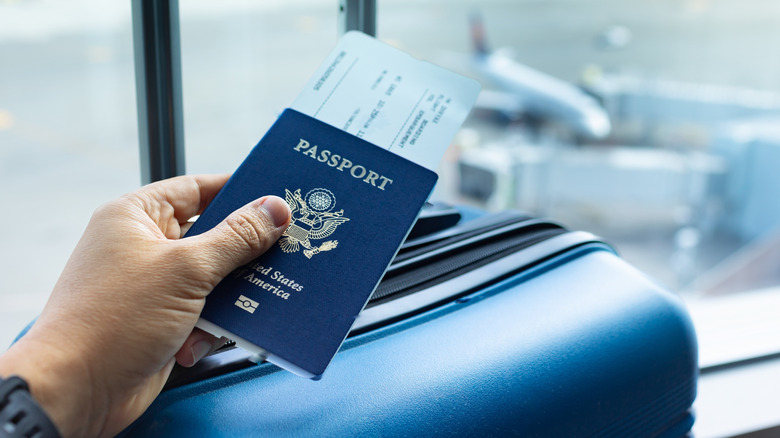 A hand holding a passport and boarding pass with a blue suitcase in the background
