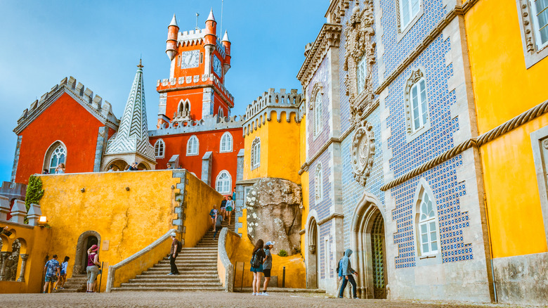 The entrance to Pena Palace, showcasing the bright colors, blue tile work, and some tourists walking a small plaza and up the stairs