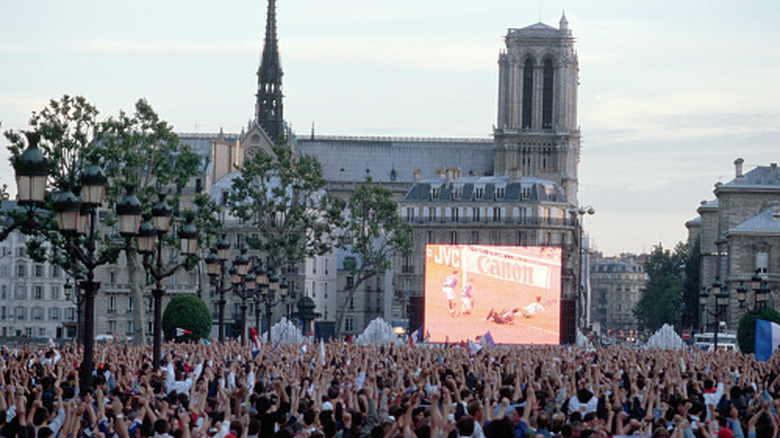 A big screen airing a football game to a crowd. European buildings in the backdrop.