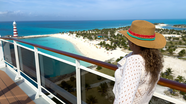 Woman in straw hat gazes from the deck of a cruise ship at a white sand beach with a lighthouse