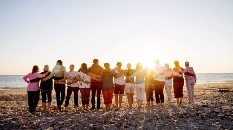 A group of tourists stand in a line facing an ocean sunset with their arms wrapped around each other