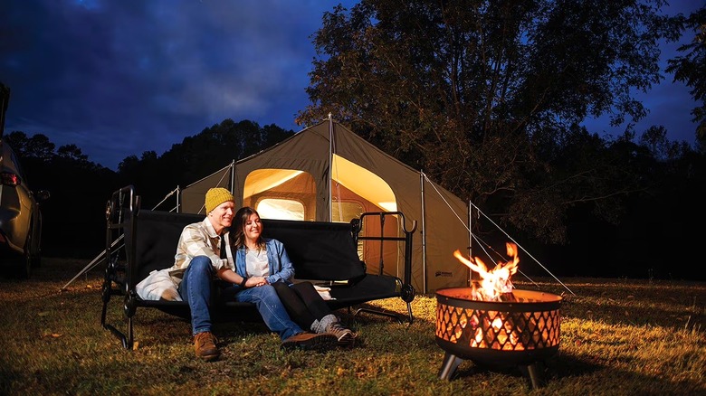 A couple sitting outside by a campfire on the bench version of the Disc-O-Bed cots with a tent in the background.