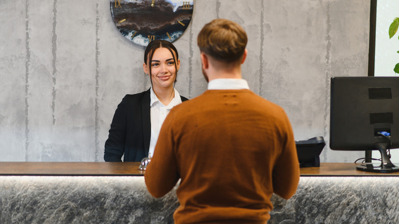 A man dressed in a brown sweater is served my a receptionist at a hotel check-in counter.