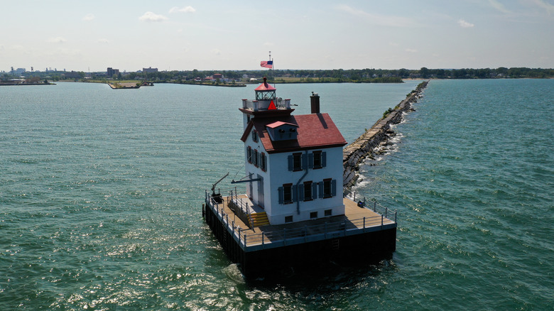 Lorain Lighthouse in the foreground, a red-roofed white building on a square platform in a blue lake, with a narrow stone path connecting it to the shoreline in the background, under a blue sky