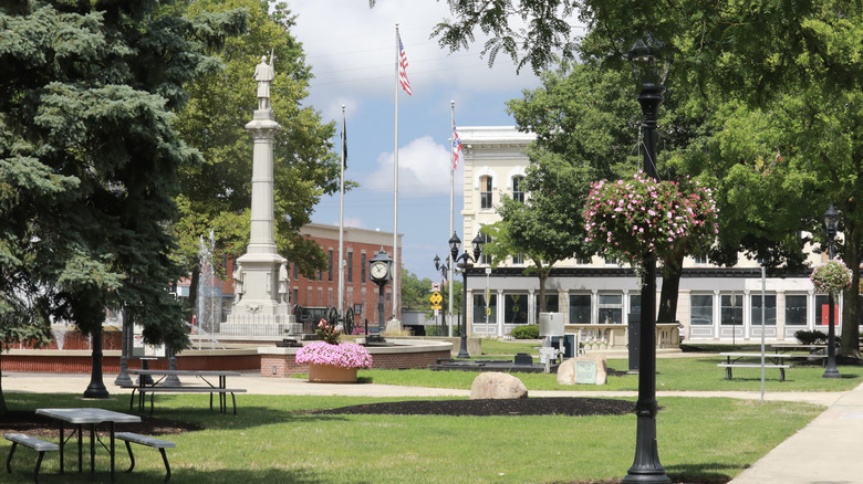 Park in Elyria, Ohio, with a gray statue-topped monument and fountain to the left and green grass with picnic tables and paths beside it, and brick and white buildings in the background, under a cloudy blue sky