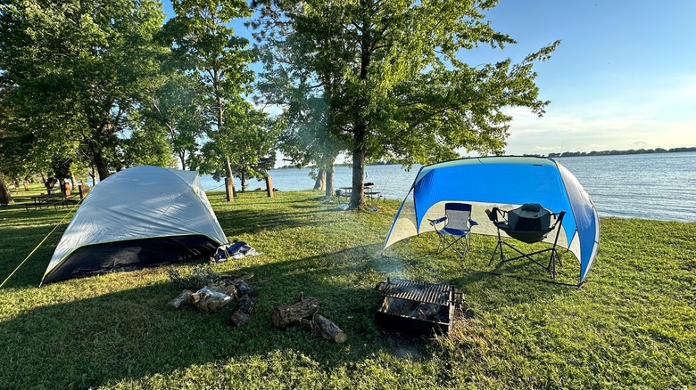 A tent and a shade awning with two chairs next to a fireplace in front of Johnson Lake with green grass, trees, blue water, and bright skies