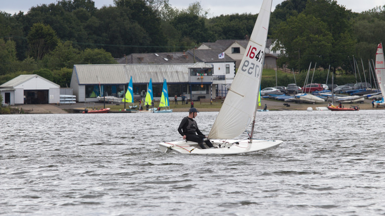 A man riding in a sailboat on Pennington Flash with the sailing club and cars behind him.
