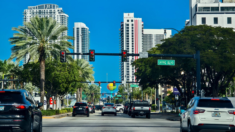 Street view of downtown Hallandale Beach with cars, trees, and condos.