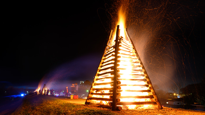 A large bonfire burns in the night while people mingle nearby and other bonfires blaze in the background.