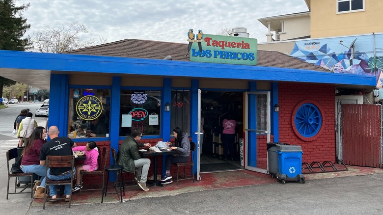 Taqueria Los Pericos' blue and orange facade and people eating at tables outside.