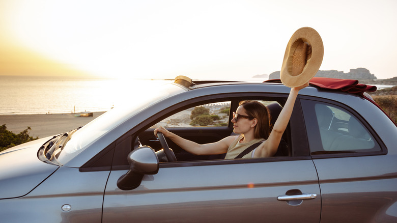 Woman driving a car along a coast and holding a hat outside the window