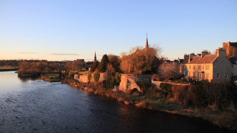 Views of an old town along the shores of a river, with trees, buildings, and stone walls at sunset