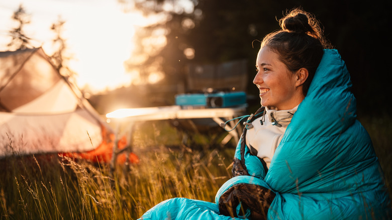 a woman sits cozy in a sleeping bag on grass, with the sunset illuminating her tent in the background