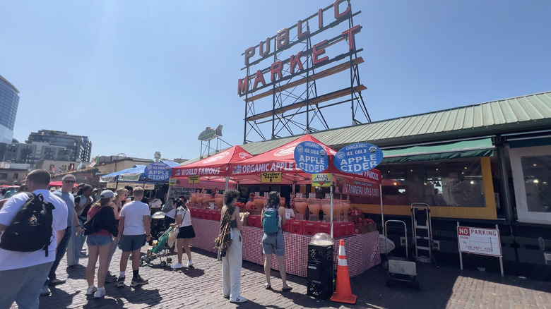 People gather around an apple cider vendor in front of Pike Place Public Market's main entrance on a sunny day in Seattle, Washington