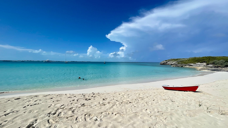 image is of a white sandy beach with calm clear blue waters; there is a red row boat sitting on the beach