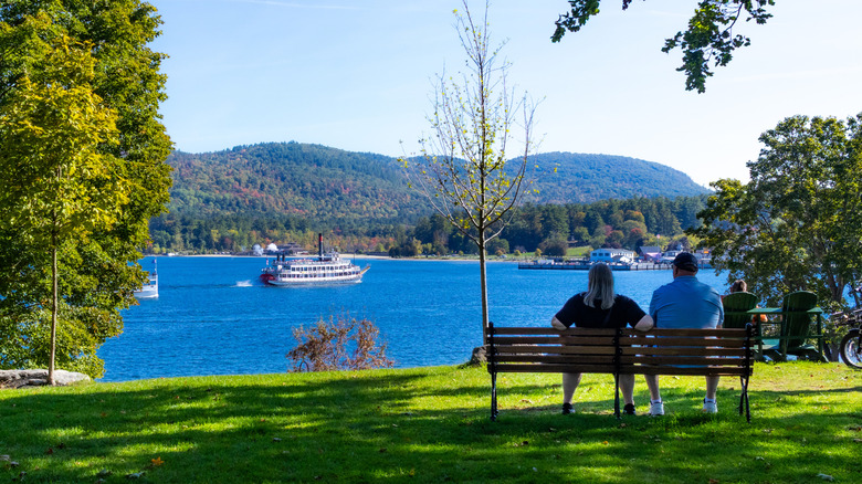 A couple sitting on a park bench on the shore of Lake George, New York, with a steamboat sailing on the lake in summer