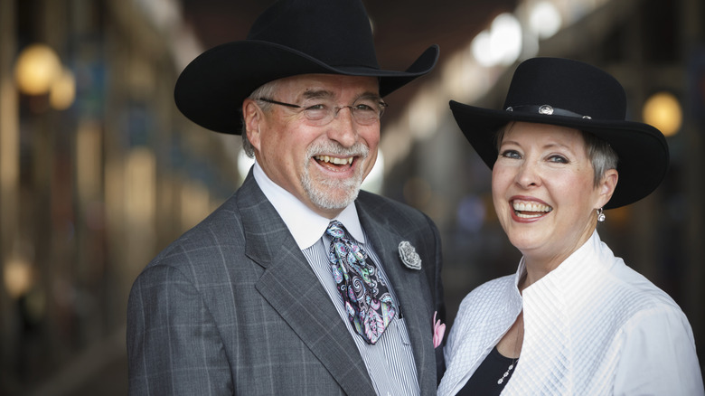 A senior couple smiles at the camera while wearing fashionable western outfits
