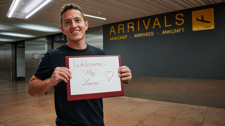 Man holding up a sign that reads "Welcome My Love" at an airport arrivals hall.