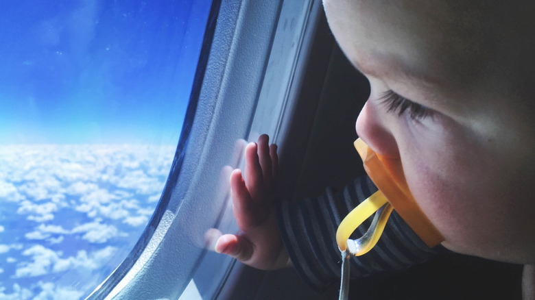 A baby sucks on a pacifier while looking out the window of an airplane.