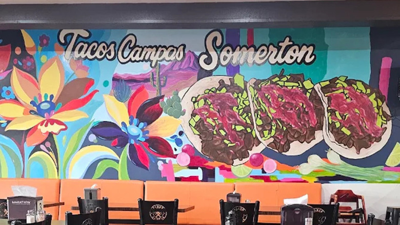 A colorful painted mural featuring multi-colored flowers and three colorful street tacos with purple salsa and "Tacos Campos Somerton" painted in white cursive with black outline