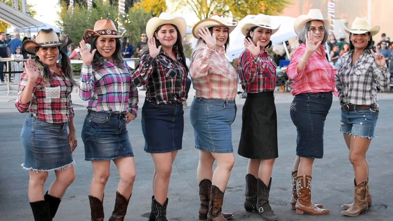 Seven women in cowboy hats and boots, flannel shirts, and denim skirts looking directly at the viewer and waving while smiling
