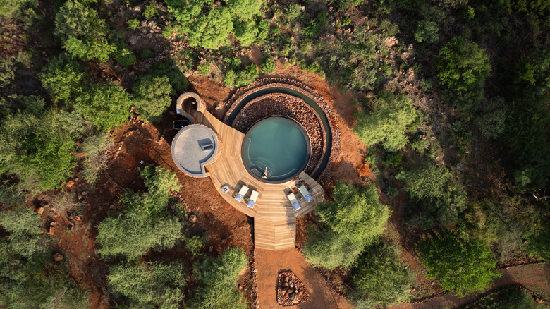 Aerial view of a secluded pool with wooden platform surrounded by trees and red rocks with a person sitting in the pool water.