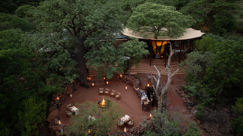 Tables and chairs surrounding a campfire with food table set up nearby, and main dining area behind, all surrounded by lush trees and red rocks.