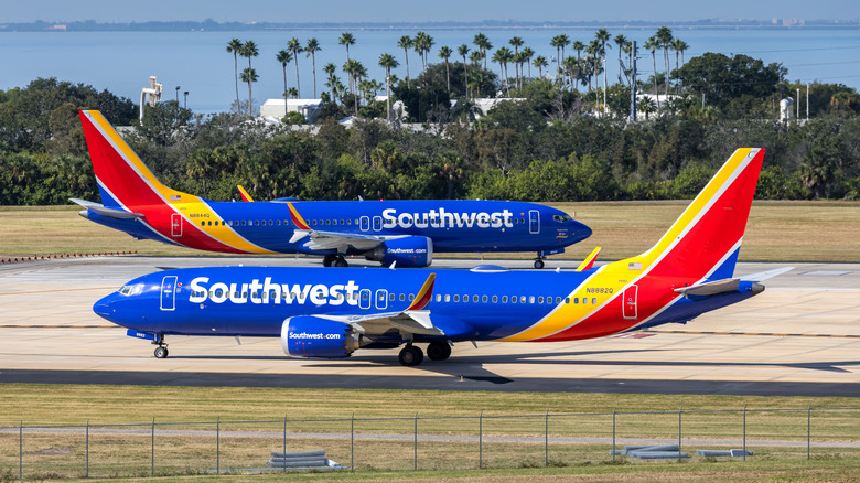 Two Southwest Airlines planes facing opposite directions, both blue with yellow and red tails, on a runway in front of trees and water under a blue sky
