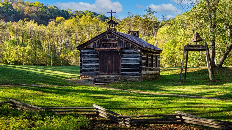 Chapel in the Pioneer Village in Beaver Creek State Park, a one-story log structure with a cross and bell on the roof and a bell on a wooden stand to the right, with a low wood fence in front of them, surrounded by grass with trees in the background under a cloudy blue sky
