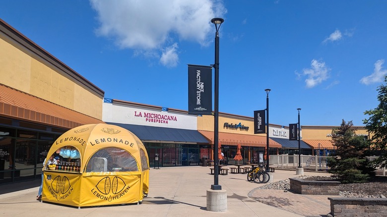 The Albertville Premium Outlets near St. Michael, with a row of stores, picnic tables on the sidewalk, and a lemonade stand