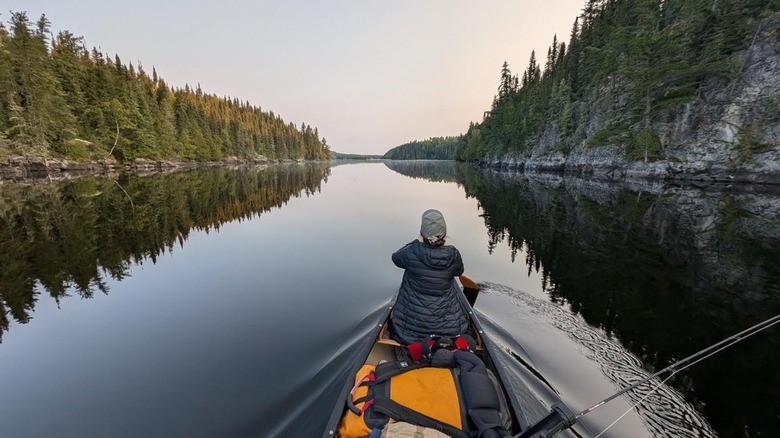 Woman paddling a canoe on a glassy lake surrounded by evergreen trees.