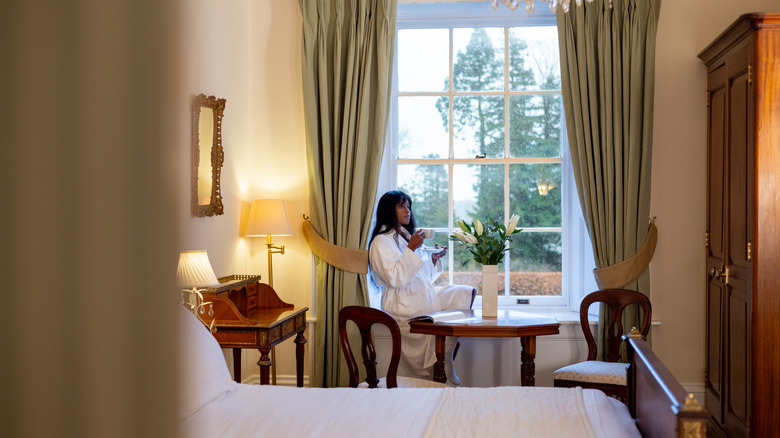 Woman sipping tea by the window in a hotel room furnished with a cabinet, writing table, and bed