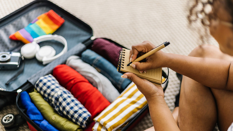 A woman writing in a notepad while sitting next to a suitcase with clothes and travel accessories.