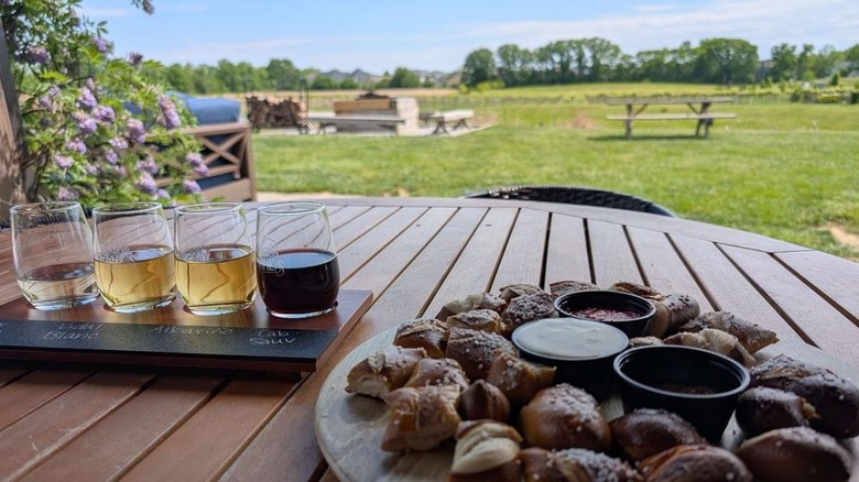 A wine flight and pretzel bites on a wooden table with scenic green fields in the background.