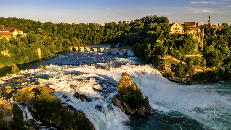 Rushing waters in the Upper Rhine river with a railway bridge and castle overlooking the falls.