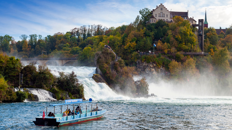 A blue tourist boat approaching the Rhine Falls in Switzerland with a castle perched above it on a cliff