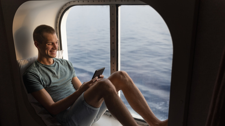 A passenger sitting in a cruise ship window while using a cell phone.