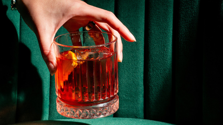 Closeup of a hand holding a Negroni cocktail in front of a green, velvet-upholstered bar booth