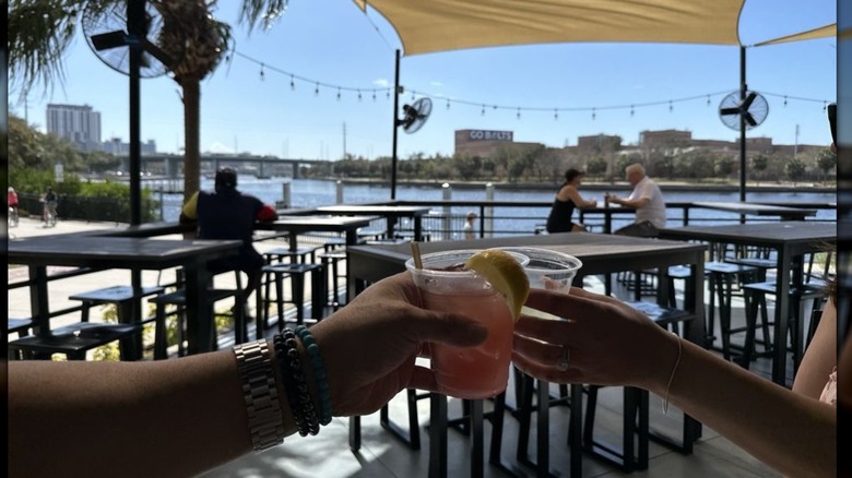 Two hands toasting cocktails on the patio of Stone's Throw with the river in the background
