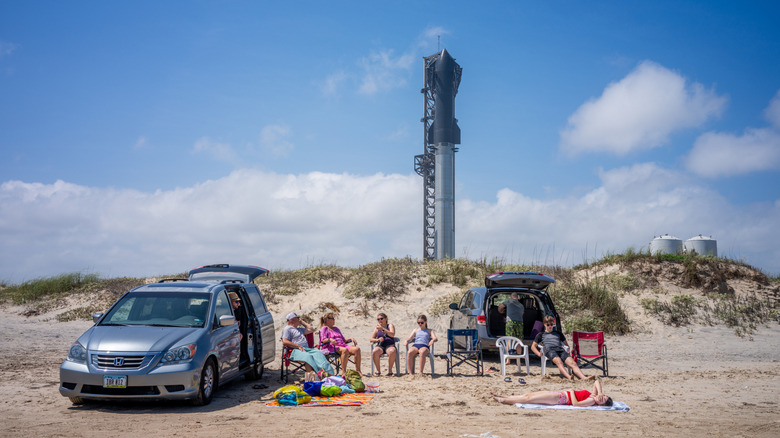 A family sit on chairs next to parked cars on a sandy dune with a rocket ship in the backdrop