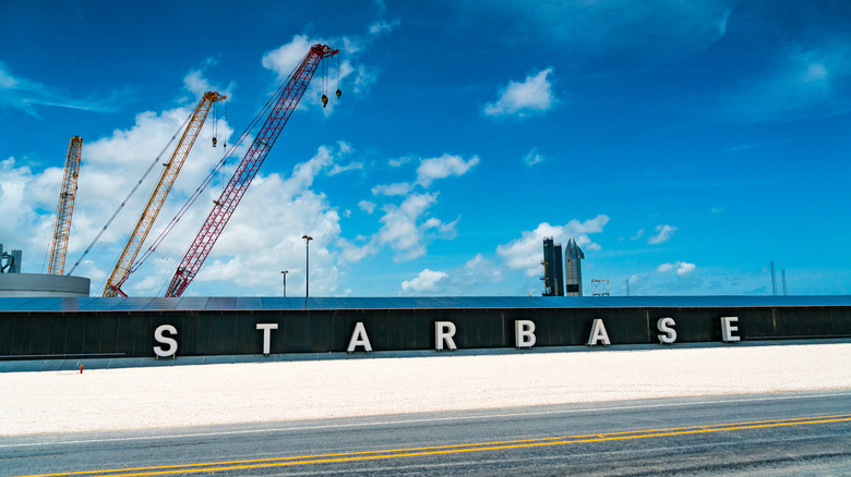 A close-up sign of Starbase on a concrete wall, with cranes and rockets against blue skies